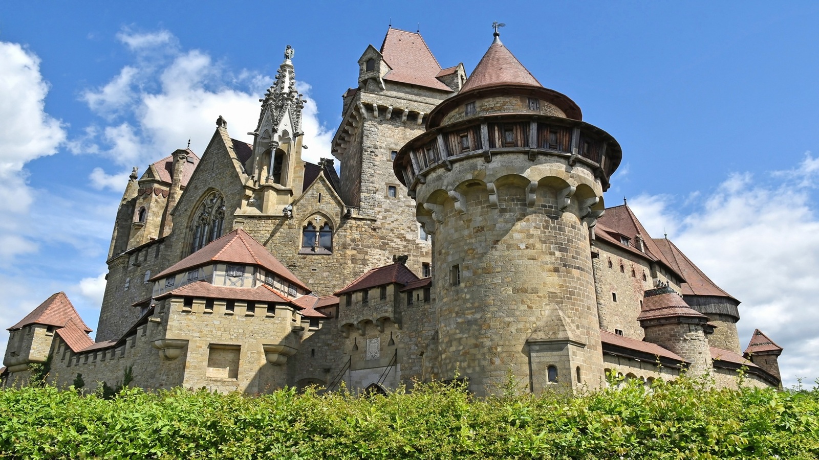 Low-angle view of a medieval stone castle with round turrets, Gothic spire, and terracotta rooftops against a blue sky