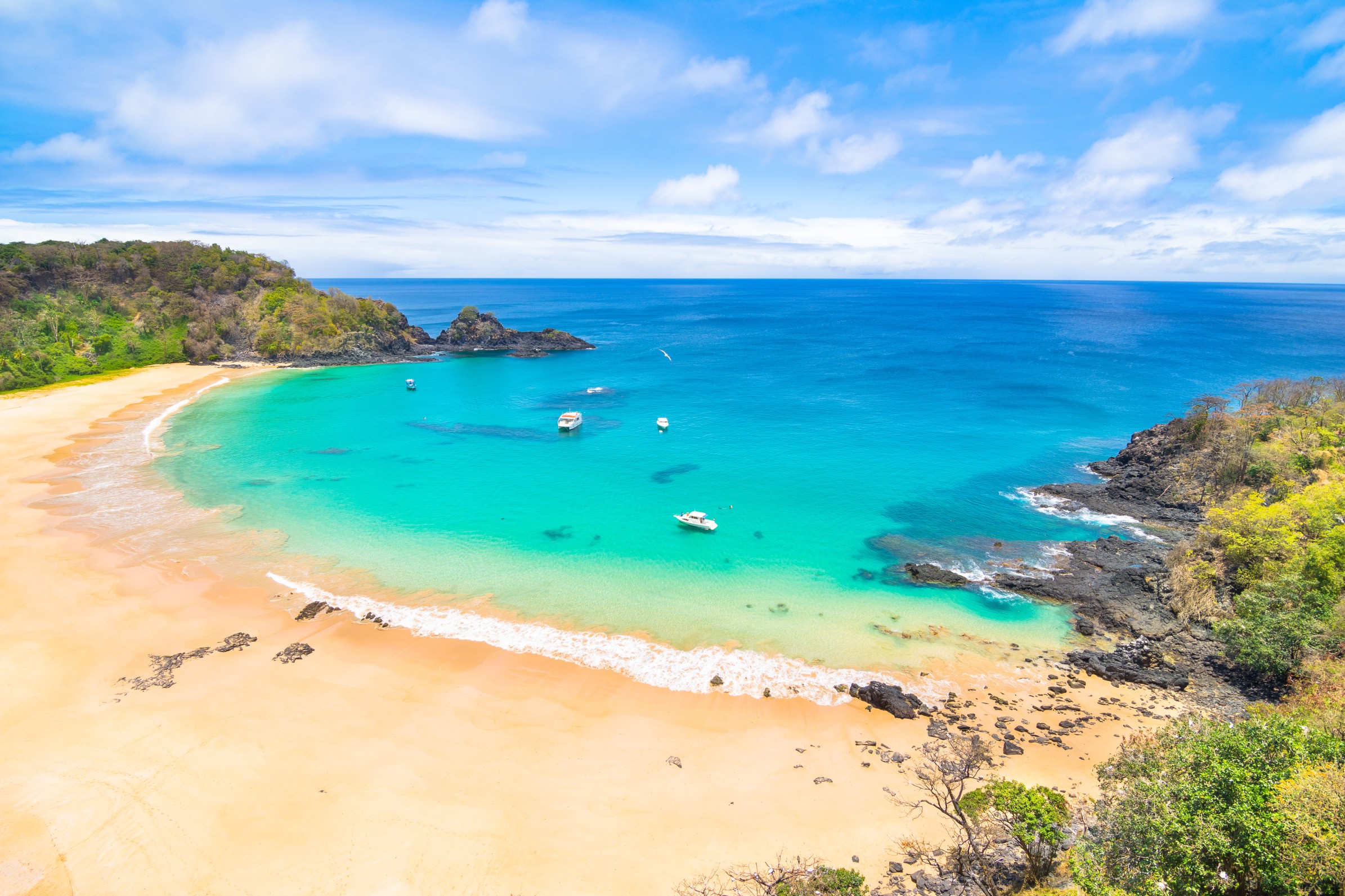 Aerial view of Baía do Sancho beach with turquoise water, golden sand, rocky cliffs, and small boats anchored in the bay at Fernando de Noronha Brazil