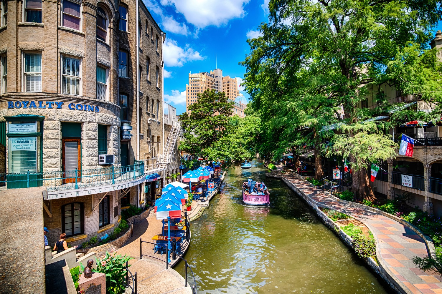 Aerial view of the San Antonio River Walk with a river cruise boat, outdoor dining patios, and lush trees on a sunny day