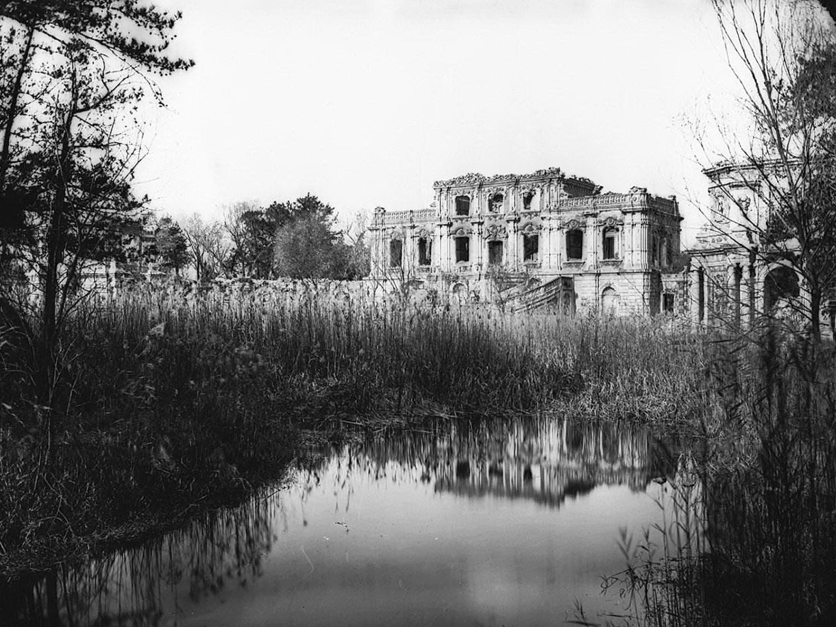 Black and white historic photograph of Old Summer Palace Beijing ruins reflected in a still pond surrounded by reeds and bare trees
