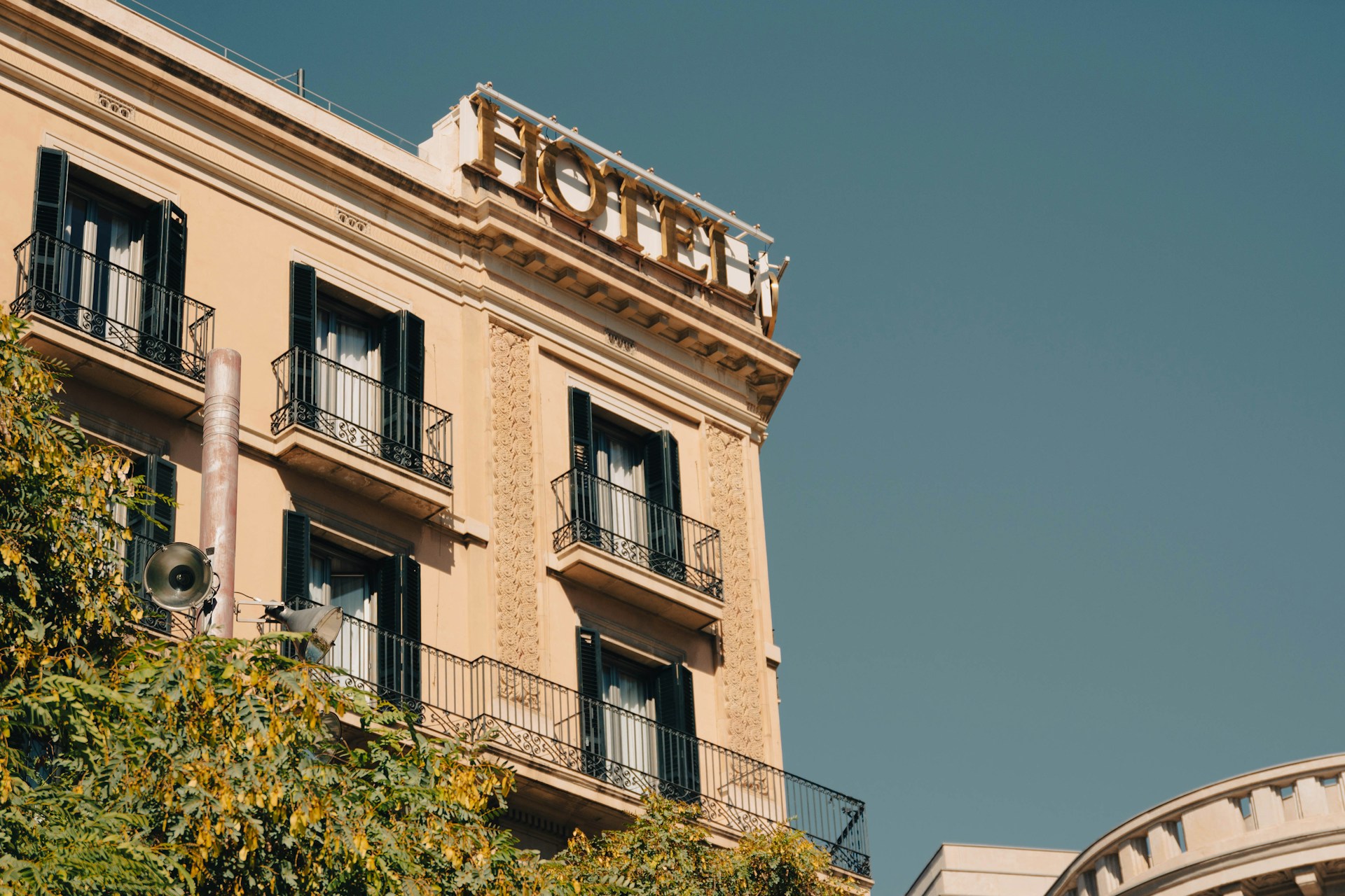 Low-angle view of a classic European hotel building with iron balconies and a gold hotel sign against a clear blue sky