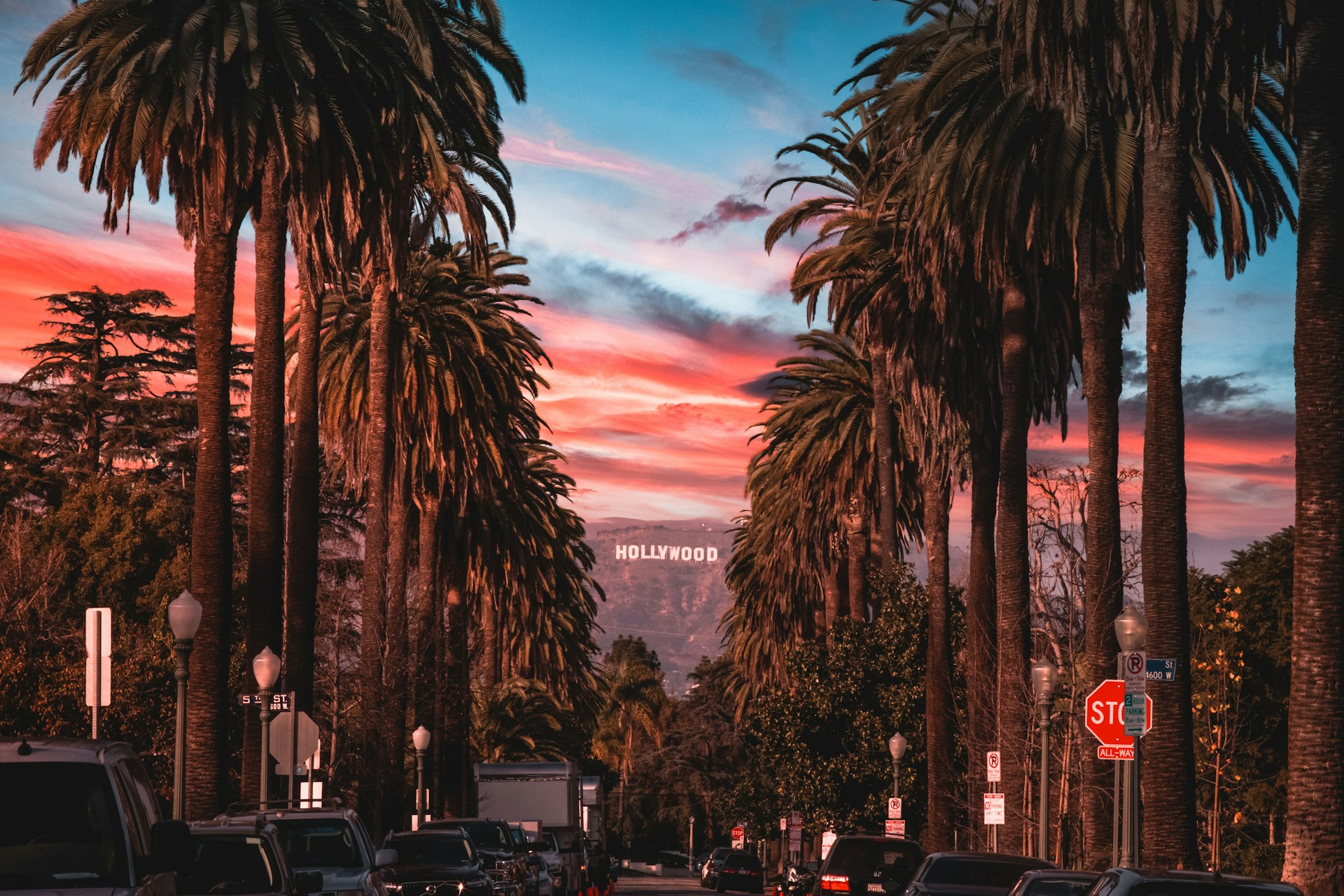 Hollywood Sign visible between tall palm trees along a Los Angeles boulevard at a vivid pink and blue sunset