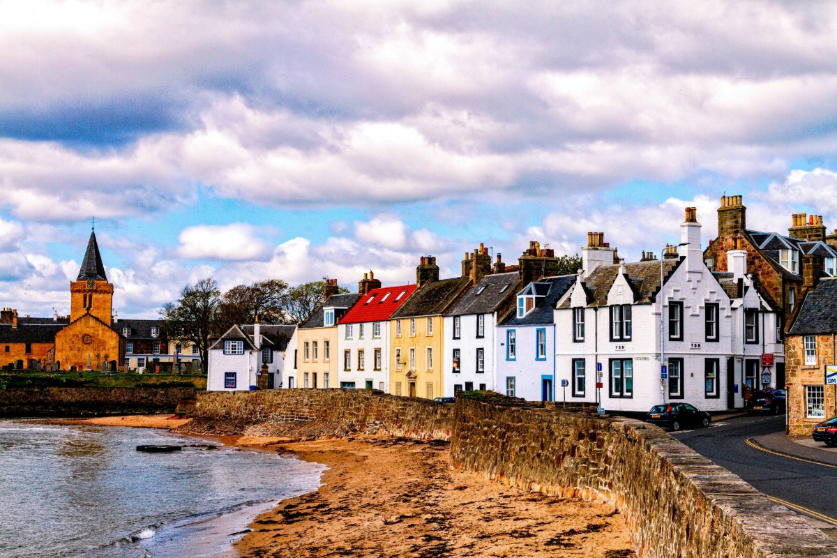 Historic Scottish coastal village with white stone buildings, a church tower, and a sandy shore under a dramatic cloudy sky