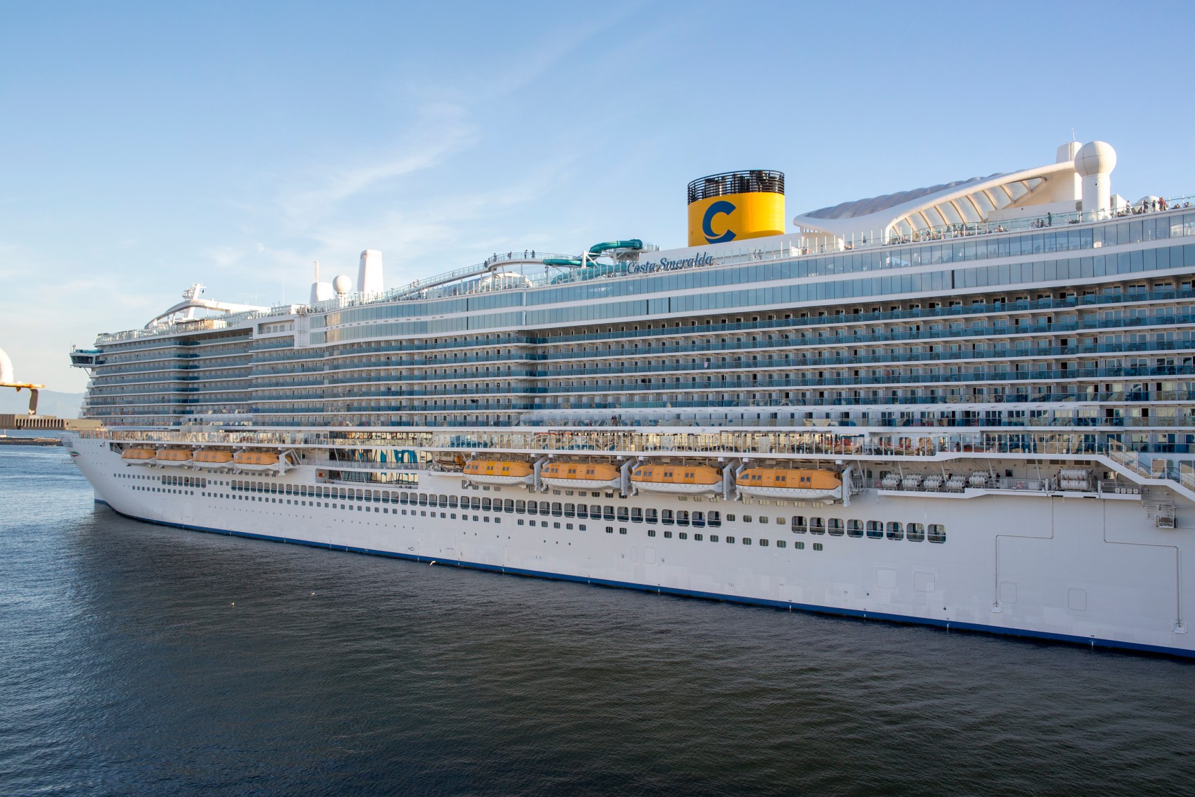 Wide angle of the Costa Smeralda cruise ship docked at port, capturing its immense scale, elegant white hull, and the iconic yellow funnel against a tranquil blue water. Naples, Italy - March, 2026