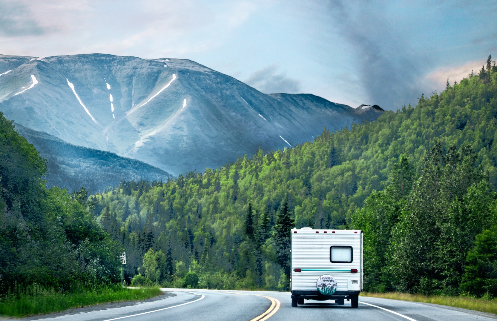 Retro camper driving on highway sucrrounded by evergreen forests and tall mountains with snow patches in distance on overcast day - Kenai Peninsula Alaska
