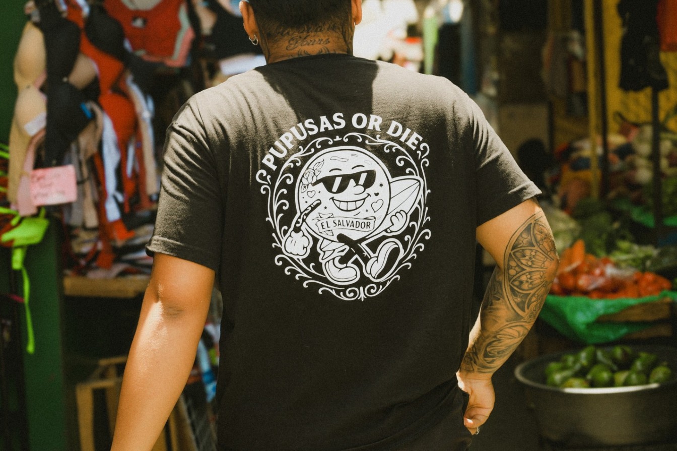 Person wearing Pupusas or Die El Salvador t-shirt standing in a colorful local market with fresh produce and clothing stalls behind