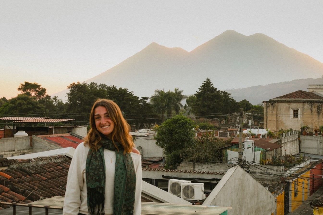 Smiling female traveler on a rooftop terrace at sunset with a Central American town and two volcanic mountain peaks in the background