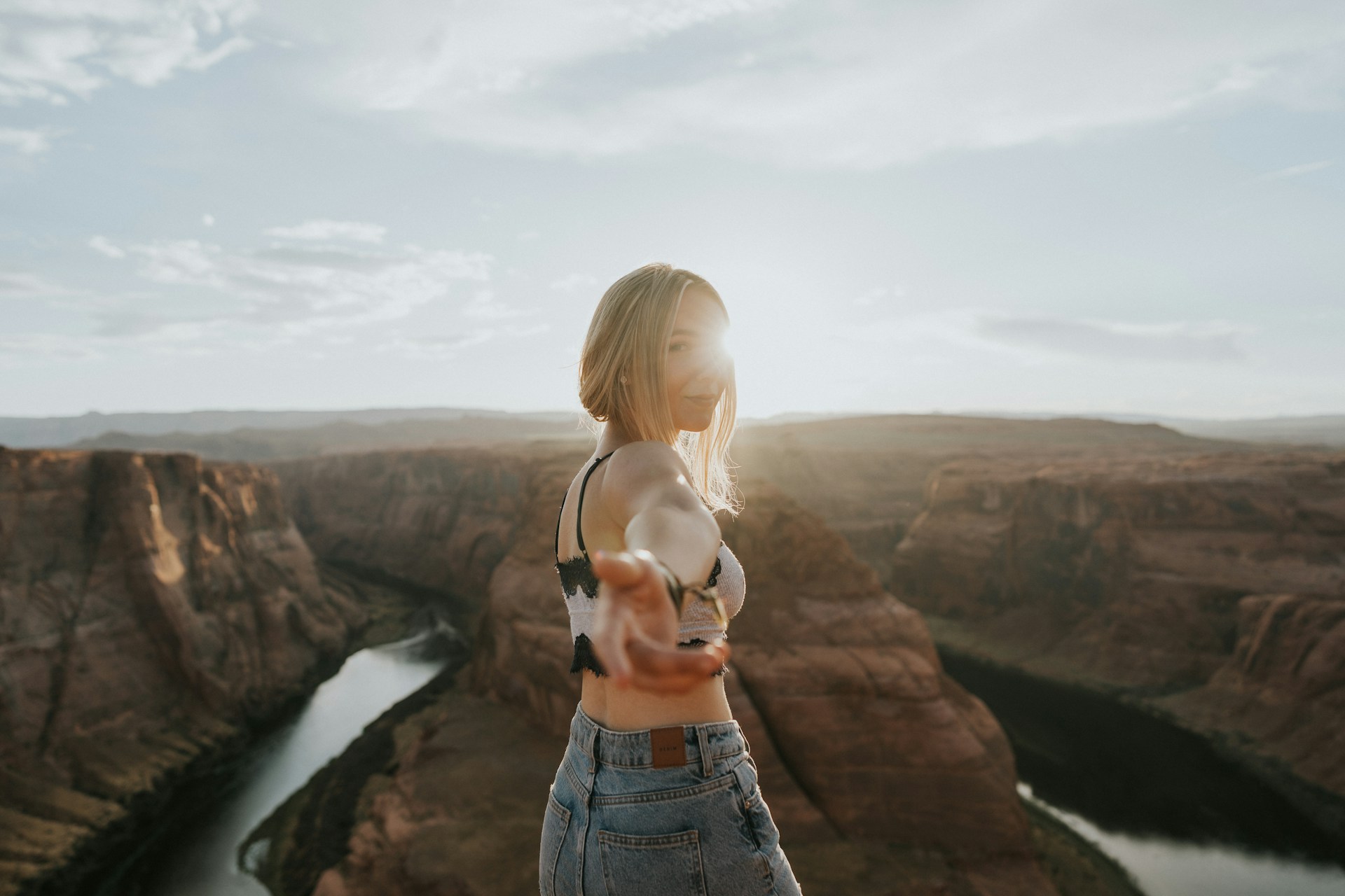 Smiling woman reaching toward camera at Horseshoe Bend canyon overlook with golden sunset light and winding river in the background